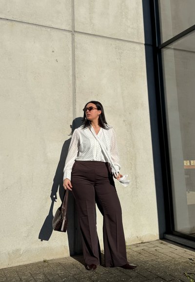 Mujer con gafas de sol, que lleva una blusa blanca de lunares y pantalones oscuros, está apoyada contra una pared de hormigón, sosteniendo un bolso marrón a la luz del sol.