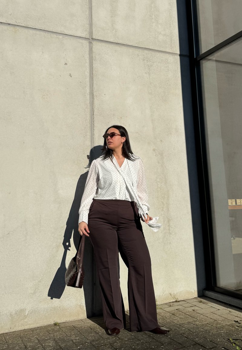 Femme avec des lunettes de soleil portant un chemisier blanc à pois et un pantalon foncé, debout contre un mur en béton, tenant un sac à main marron au soleil.