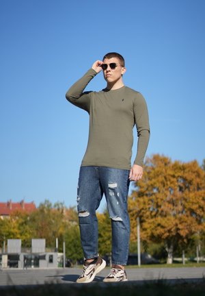 Camisa de manga larga verde oliva, jeans azules desgastados con desgarros y zapatillas deportivas marrones. Accesorio: gafas de sol negras.