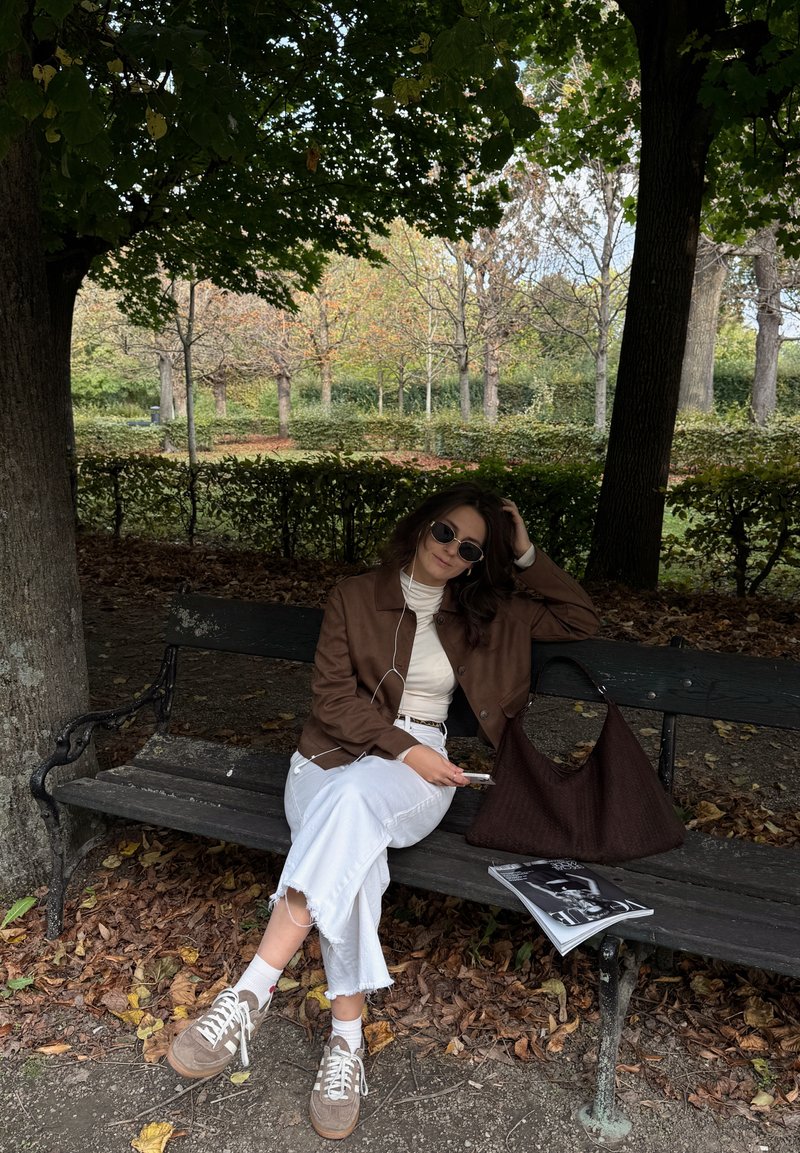 Giubbotto di pelle marrone sopra un dolcevita bianco, abbinato a jeans bianchi a vita alta e sneakers. Borsa intrecciata marrone scuro e riviste su una panchina.
