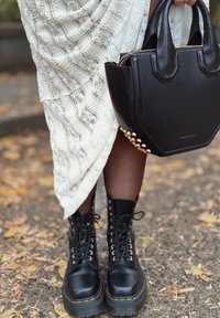 Zwarte leren enkellaarzen met ronde neus, vetersluiting en dikke zolen. Zwarte handtas met gouden studs en ritssluiting. Witte textuur jurk.