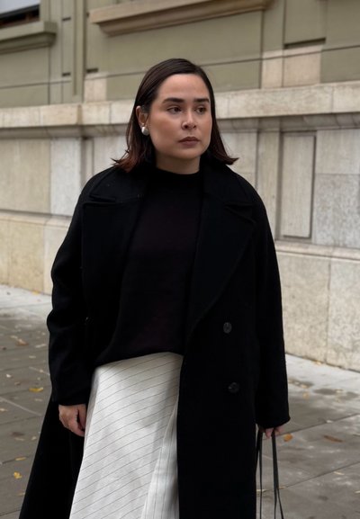 Abrigo de mezcla de lana negra con grandes botones, llevado sobre una blusa negra y una falda crema con rayas grises. El fondo es una pared de piedra.