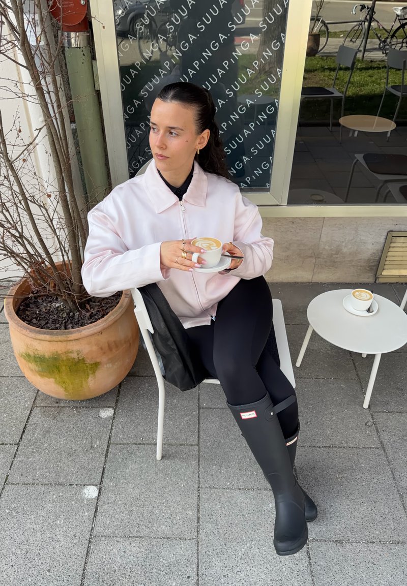 Kvinne i lys jakke og svarte støvler sitter utendørs på hvit stol, holder en kaffekopp, ved siden av et lite rundt bord med en annen kopp.