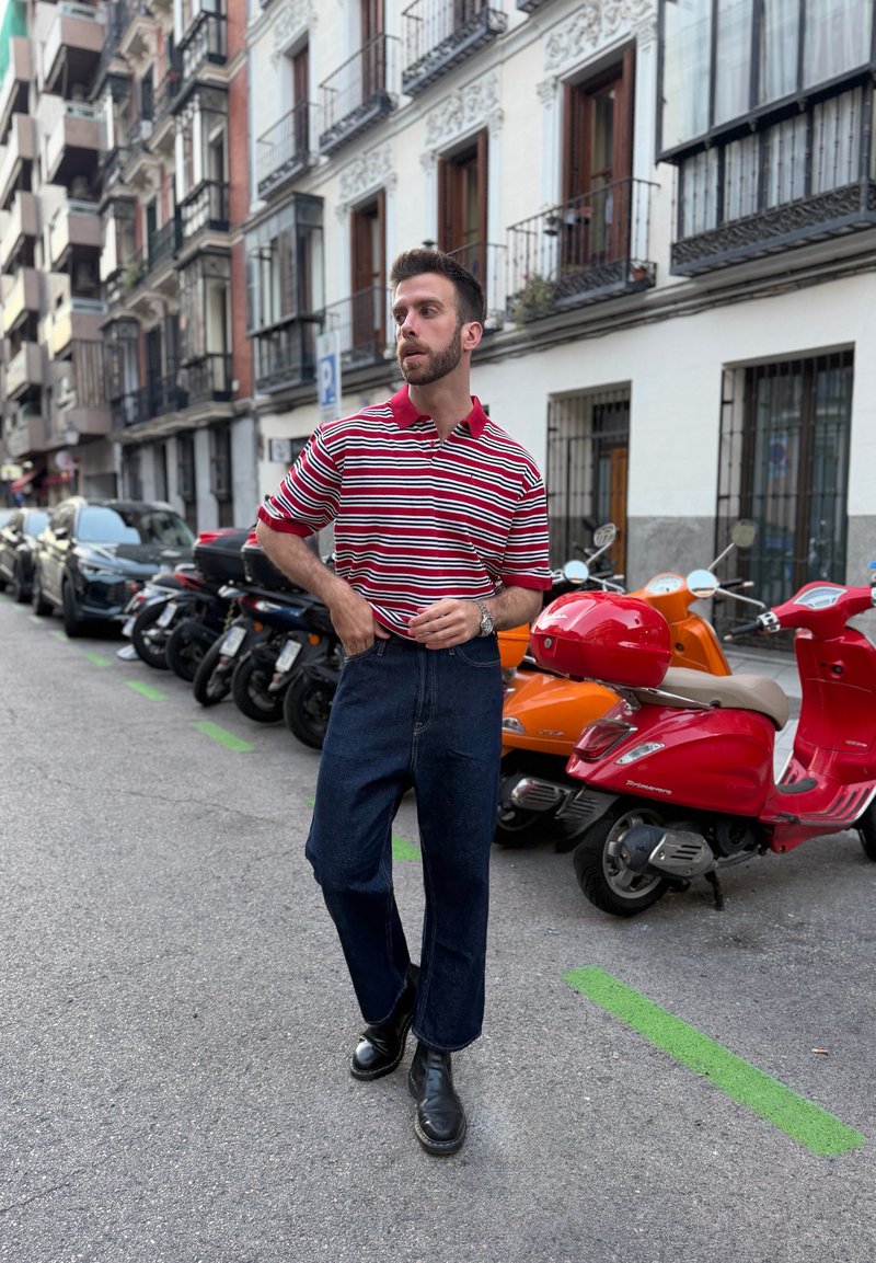 Homme en chemise rayée rouge et blanche et jean foncé debout dans une rue avec des scooters et des voitures garés près des immeubles résidentiels.