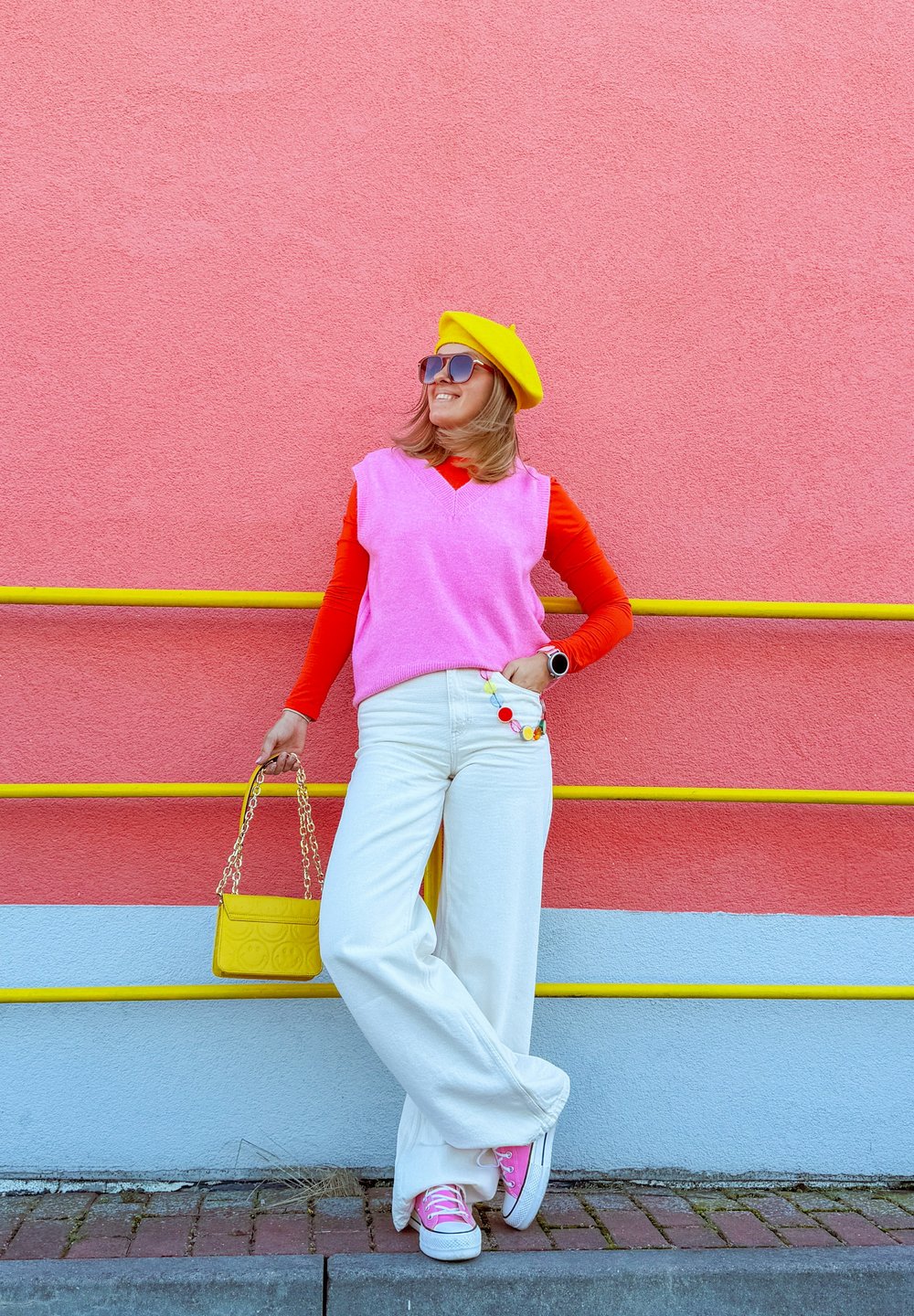 Roze mouwloze trui over een rode langemouwen shirt, witte brede broek, roze sneakers, gele baret, geelkleurige tas en oversized zonnebril.