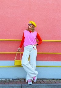 Roze mouwloze trui over een rode langemouwen shirt, witte brede broek, roze sneakers, gele baret, geelkleurige tas en oversized zonnebril.