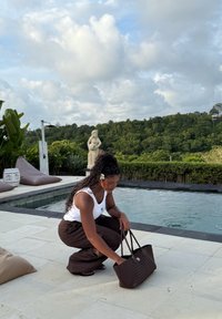 Femme avec une fleur dans les cheveux accroupie au bord de la piscine, plongeant la main dans un grand sac tissé, entourée de verdure et sous un ciel nuageux.