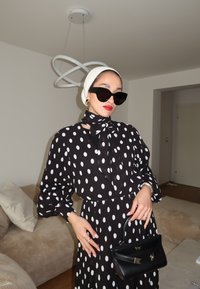 Vestido preto com bolinhas brancas, com um lenço amarrado no pescoço, óculos de sol oversized pretos e uma pequena mala preta. Tecido suave, mangas longas e cintura ajustada.