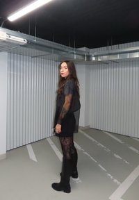 Crna prozirna majica s dugim rukavima, uparena s crnom mini suknjom i tajicama s čipkastim uzorkom. Crne čizme do koljena upotpunjuju izgled.