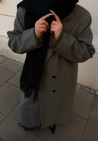 Grijze wollen overjas met grote zwarte knopen, gecombineerd met een zwarte sjaal met franjes. Lange, puntige zwarte laarzen, handen die een klein voorwerp vasthouden.