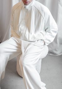 Weißer Reißverschlussjacke mit vertikalen Falten, kombiniert mit lockeren, weißen Hosen. Glatter, leichter Stoff; minimalistisches Design mit einem subtilen Schimmer.