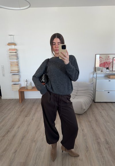 Suéter gris de punto, pantalones de piernas anchas marrón con rayas, botas de tacón marrón claro y un pequeño bolso negro. Fondo minimalista con libros y muebles.
