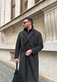 Homme portant un long manteau sombre et des lunettes de soleil, tenant un sac noir, debout sur le trottoir près d'un bâtiment en pierre beige avec des fenêtres.
