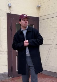 Νέος άνδρας με γκρι πουκάμισο, σκούρο παλτό, γκρι παντελόνι και μπορντό καπέλο στέκεται κοντά στην είσοδο ενός κτηρίου με μπεζ τούβλα.