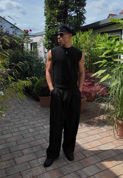 Top negro sin mangas combinado con pantalones negros holgados, complementado con zapatos negros. Rodeado de exuberante vegetación y pavimento de ladrillo.