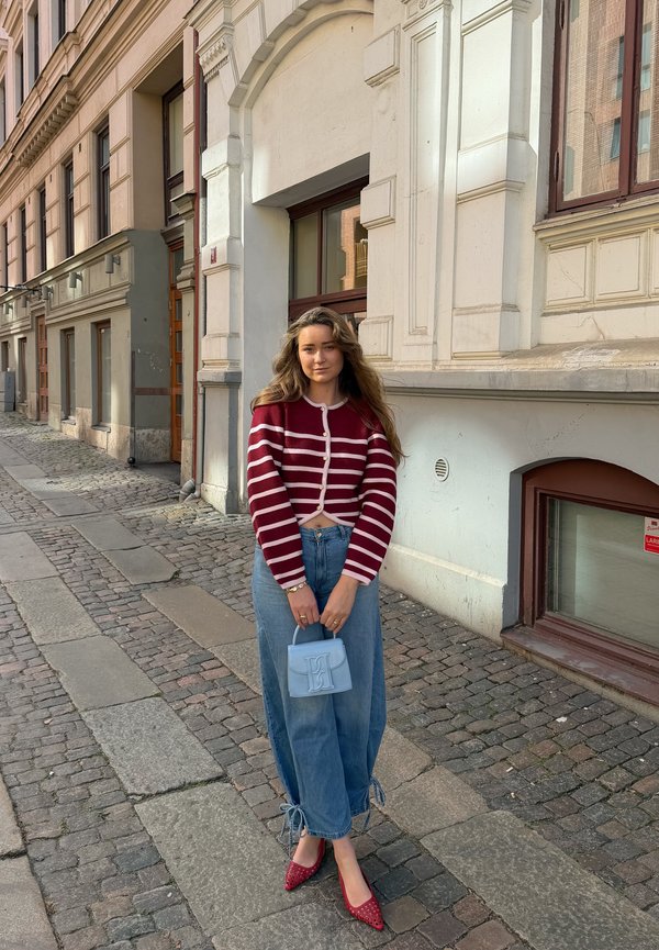 Joven mujer de pie en una calle empedrada con un suéter de rayas rojas, vaqueros anchos azules, zapatos rojos y sosteniendo un pequeño bolso azul claro.
