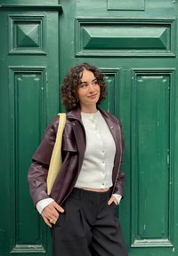 Bordeauxrode leren jas met een oversized pasvorm, crèmekleurige trui met knopen, zwarte broek en een lichtbeige tas tegen een groene deur.