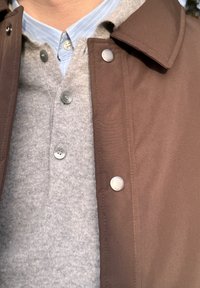 Geacă exterioară maro cu nasturi cu capse peste un pulover gri deschis. Puloverul are o clapetă cu nasturi gri și un guler cu dungi albastre deschis.