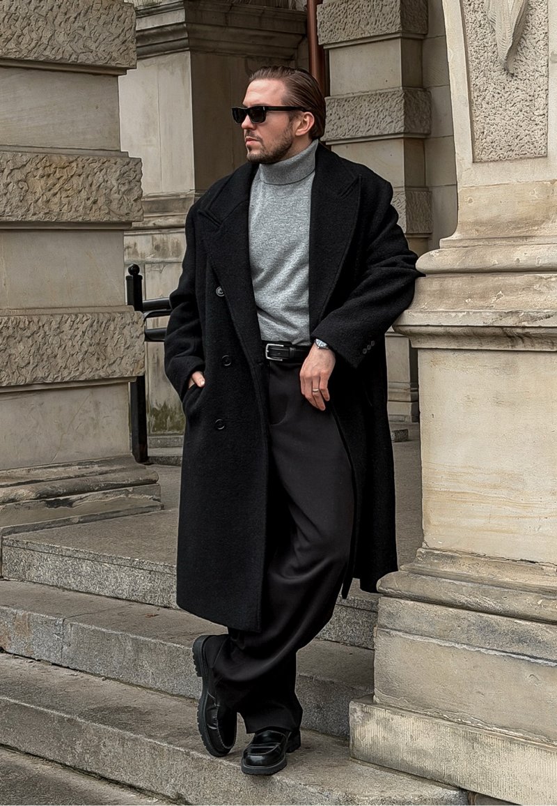 Manteau long en laine noire sur un col roulé gris, pantalon noir et bottes noires brillantes. Debout sur des marches en pierre avec un fond texturé.