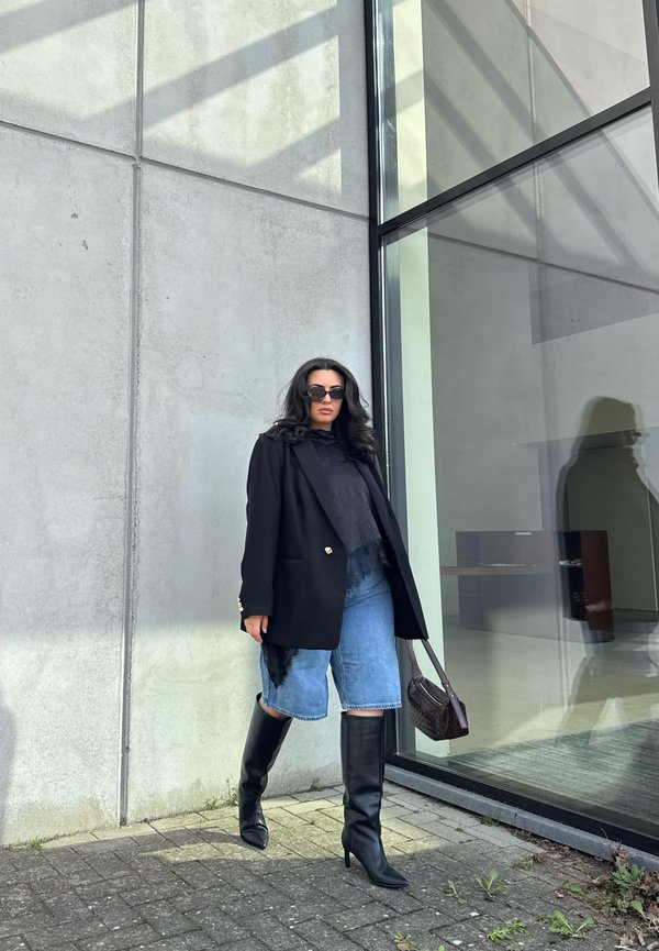 Femeie purtând blazer negru, ochelari de soare, cizme negre peste genunchi și pantaloni scurți din denim, stând lângă o clădire de sticlă și beton.