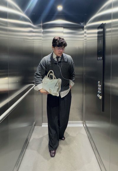 Hombre con gafas y auriculares, sosteniendo un bolso blanco, de pie dentro de un ascensor de acero inoxidable con el panel de control iluminado en el piso 3.