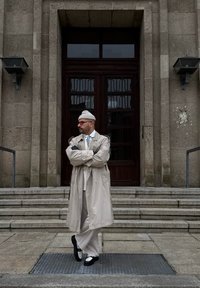 Μπεζ παλτό τρέντς πάνω από ένα λευκό ρούχο, μαύρα και άσπρα παπούτσια και ένα ανοιχτό beanie. Στέκεται σε σκαλοπάτια με σχέδιο στον τοίχο πίσω.