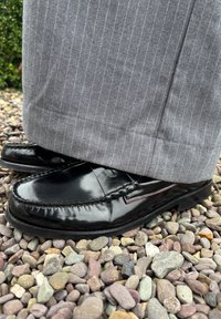 Mocasines negros brillantes con un diseño cosido, llevados con pantalones a rayas grises, descansando sobre una cama de guijarros.