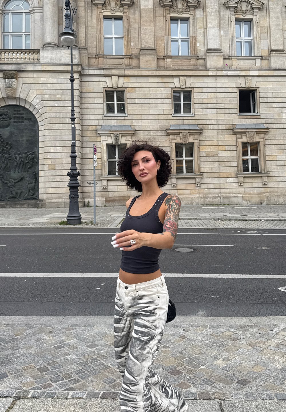 Top senza maniche nero con accenti di borchie, pantaloni bianchi con motivo astratto grigio, capelli lunghi e una piccola borsa nera. Sfondo urbano di pietra.