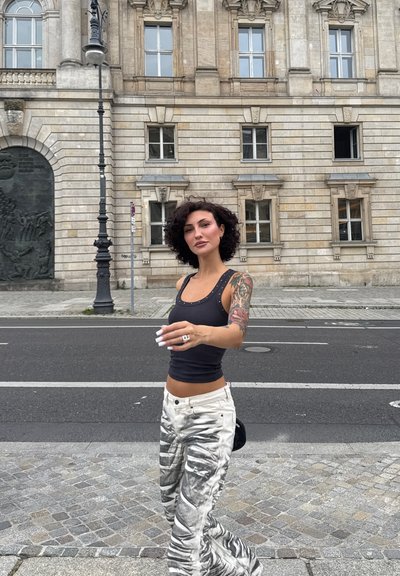 Top negro sin mangas con acentos de tachuelas, pantalones blancos con patrón abstracto gris, cabello largo y una pequeña bolsa negra. Fondo de piedra urbana.