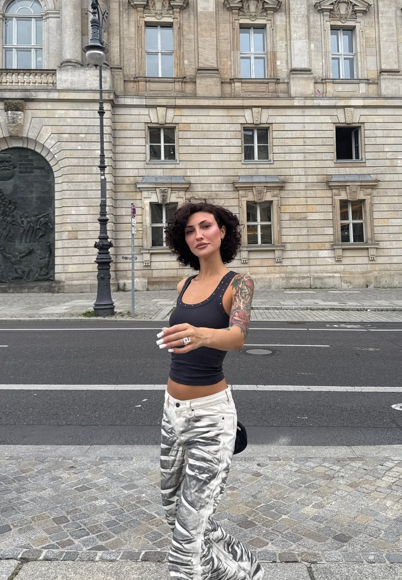Top nero senza maniche con dettagli di borchie, pantaloni bianchi con un motivo astratto grigio, capelli lunghi e una borsa nera piccola. Sfondo urbano in pietra.