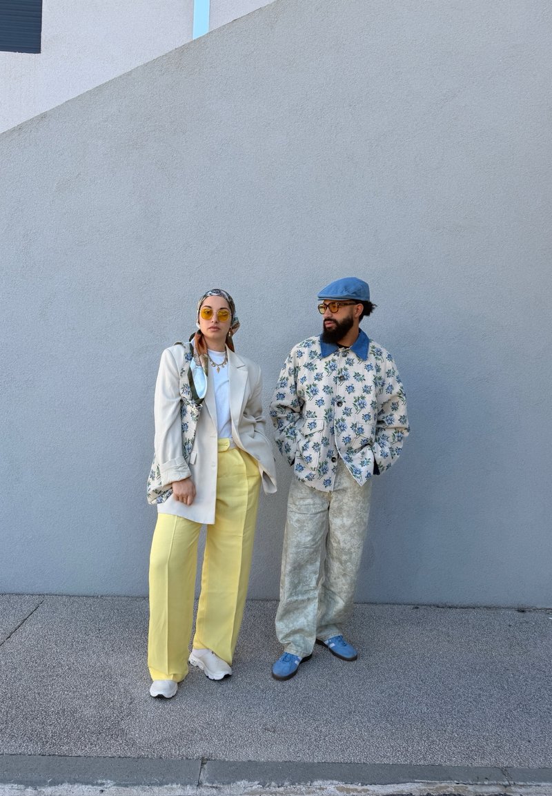 Deux adultes élégants se tiennent contre un mur gris : l'un porte un pantalon jaune et un blazer blanc, l'autre porte une veste et un pantalon à motifs avec un chapeau bleu.