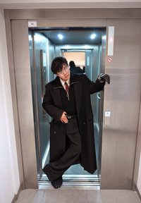 Personne en manteau noir, gants et lunettes se tient décontractée dans l'embrasure d'un ascenseur ouvert, posant avec une jambe croisée et la main sur la taille.