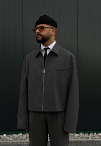 Γκρίζο κοντό μπουφάν με φερμουάρ στο μπροστινό μέρος, μακρύ μανίκι και τσέπη στο στήθος. Φορεμένο πάνω από ένα λευκό πουκάμισο και μαύρη γραβάτα, με μαύρο σκουφάκι και γυαλιά ηλίου.