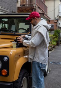 Mees punase mütsi, valge särgi ja teksadega seisab kollase vintage maastikuauto kõrval, asetades musta kotti selle kapoti peale, mille kõrval on jook.