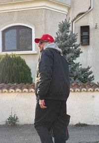 Veste bomber noire portée sur un sweat à capuche gris, pantalon noir et casquette rouge. En arrière-plan, de la verdure et un mur beige avec des tuiles décoratives sur le toit.