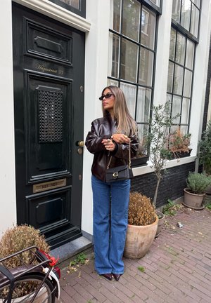 Chaqueta de cuero marrón, vaqueros anchos azules y un pequeño bolso negro. De pie junto a una puerta negra con detalles decorativos y plantas cercanas.