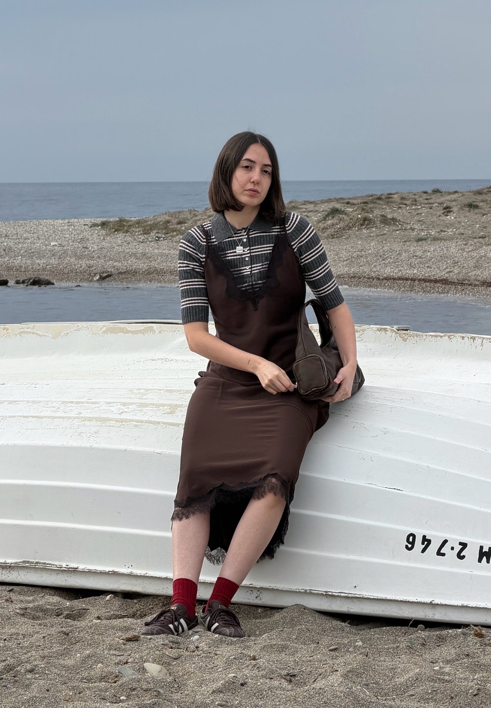 Νεαρή γυναίκα με ριγέ πουκάμισο και καφέ φόρεμα κάθεται σε αναποδογυρισμένη λευκή βάρκα σε βοτσαλωτή παραλία δίπλα σε ήρεμη θάλασσα, κρατώντας καφέ τσάντα.