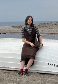 Νεαρή γυναίκα με ριγέ πουκάμισο και καφέ φόρεμα κάθεται σε αναποδογυρισμένη λευκή βάρκα σε βοτσαλωτή παραλία δίπλα σε ήρεμη θάλασσα, κρατώντας καφέ τσάντα.