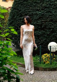 Σατέν βραδινό φόρεμα με λεπτές τιράντες, κρεμ χρώματος με αφηρημένα μπεζ σχέδια. Συνδυάζεται με μια μικρή ασημί τσάντα και κομψά υποδήματα.