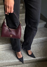 Sac en cuir bordeaux avec une forme incurvée et des accessoires en or, tenu par une main, associé à un jean noir et des talons noirs pointus sur des marches d'escalier.