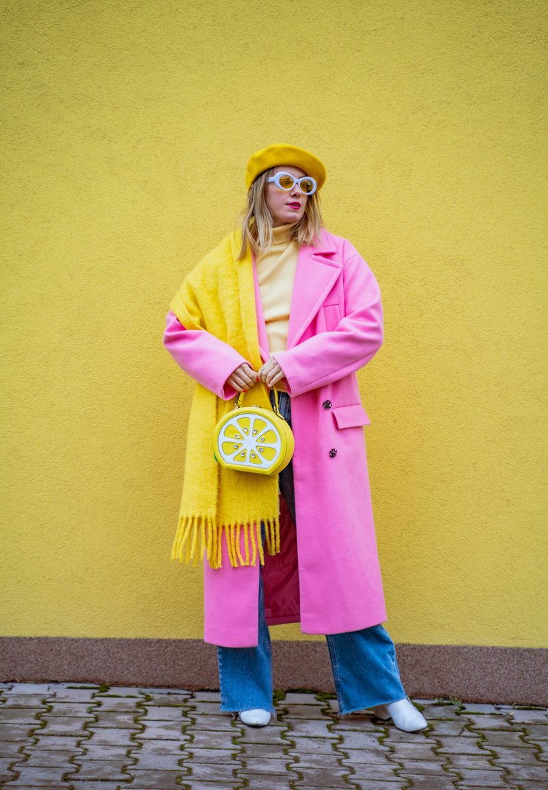 Kvinna står mot en gul vägg iklädd en ljusrosa rock, gul halsduk och basker, vita solglasögon, blå jeans, vita skor, håller en väska formad som en citron.