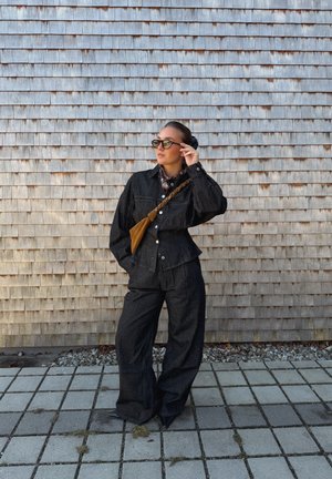 Mujer con pantalones anchos negros y chaqueta, bolso cruzado marrón, gafas de sol y bufanda, de pie frente a una pared de tejas de madera.