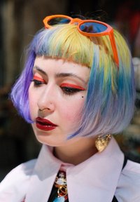 Une coupe de cheveux bob courte, pastel, dans les tons bleu, jaune et violet. Des lunettes de soleil orange vif, du maquillage pour les yeux rouge audacieux, et une couleur de lèvres rouge foncé brillante.
