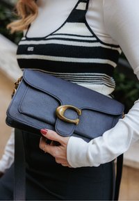 Geantă crossbody din piele bleumarin cu finisaj texturat, având o închizătoare proeminentă din metal auriu și o curea detașabilă.