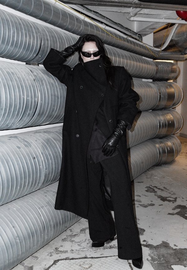 Persona con cappotto nero, guanti e occhiali da sole che si appoggia a grandi condotti di ventilazione industriale in un corridoio seminterrato scarsamente illuminato.