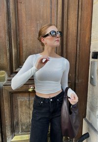 Wit langmouw cropped top, zwarte high-waisted jeans en een bruine leren schoudertas. Zwarte oversized zonnebril maakt de look compleet.