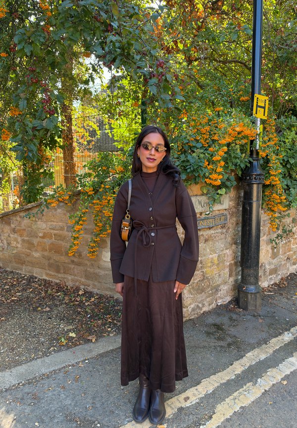 Mujer con gafas de sol, chaqueta marrón, falda larga y botas, de pie en una esquina de la calle con muro de piedra y bayas naranjas en la vegetación detrás de ella.