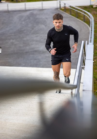 Hombre con tatuajes vestido con ropa deportiva negra subiendo escaleras de concreto al aire libre junto a una barandilla de metal.