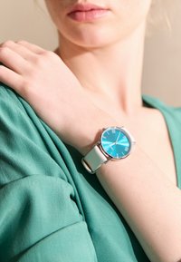 Runde Uhr mit grünblauem Ziffernblatt, silberner Hardware und weißem Lederarmband. Getragen an einem hellgrünen Ärmel. Minimalistisches Design.