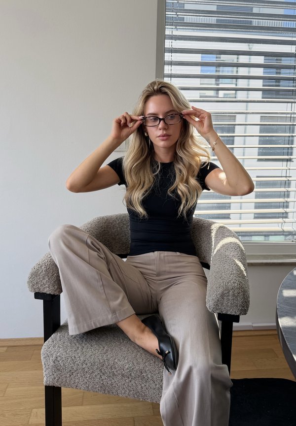 Femme aux longs cheveux blonds portant des lunettes, une chemise noire et un pantalon beige, assise en tailleur sur un fauteuil texturé près d'une fenêtre avec des stores.